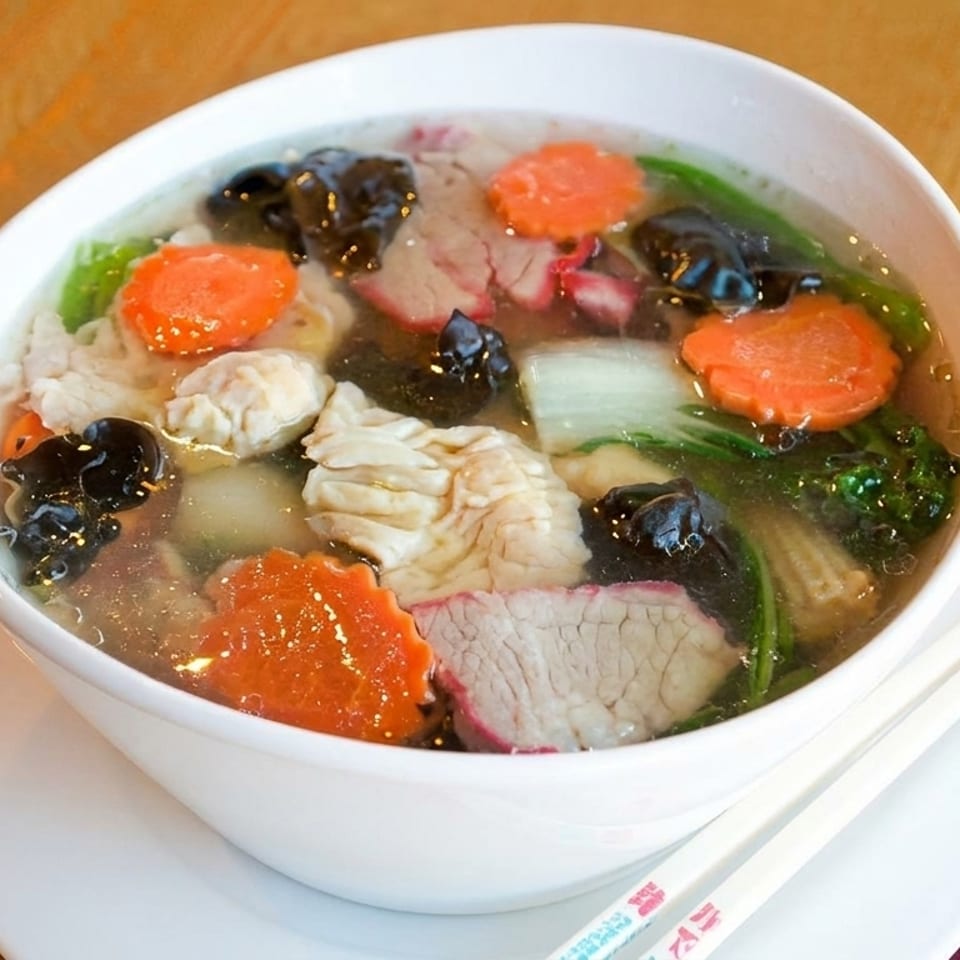 A bowl of clear soup with sliced carrots, bok choy, mushrooms, and various meats, with chopsticks.