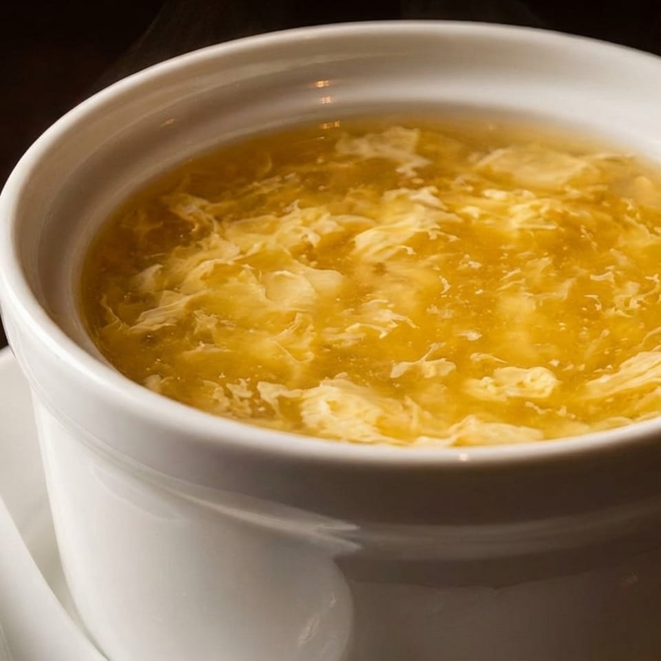 Steaming hot egg drop soup in a white bowl with a white spoon on a dark background.